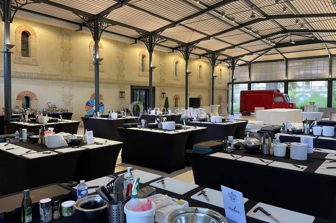 Salle aménagée avec des tables noires équipées de matériel de cuisine, sous une verrière avec un camion rouge en arrière…
