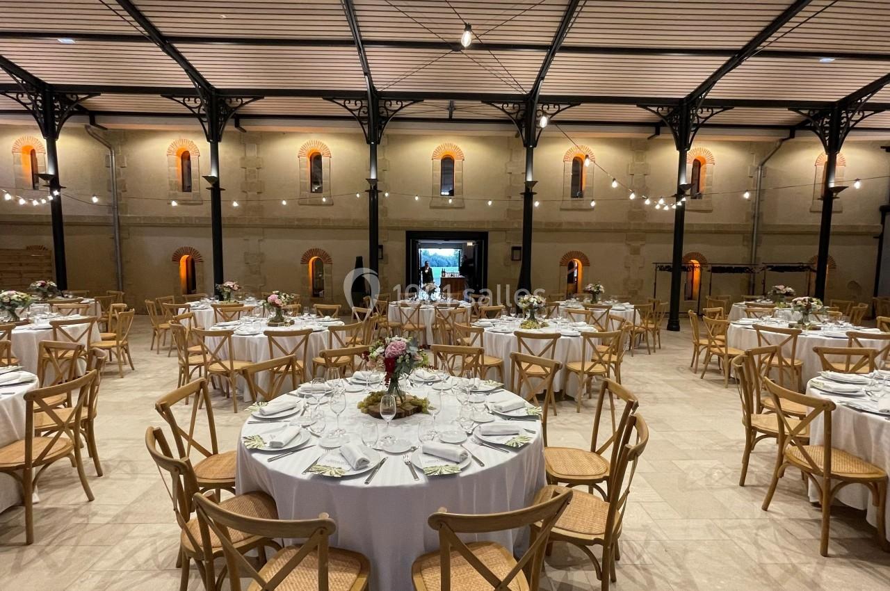 Salle de réception avec tables rondes dressées, chaises en bois et éclairage suspendu dans un cadre aux murs en pierre.