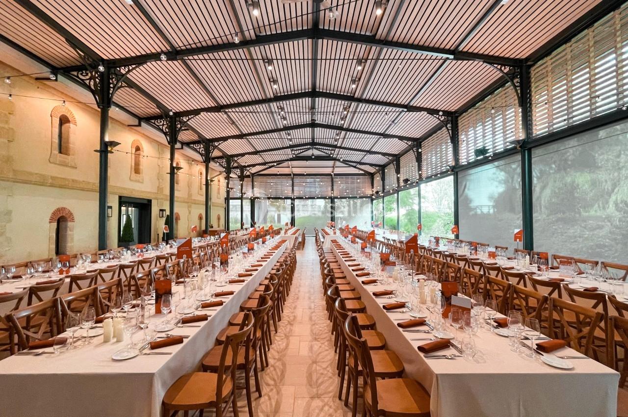 Grande salle de réception avec de longues tables dressées, chaises en bois et lumière naturelle entrant par les fenêtres.