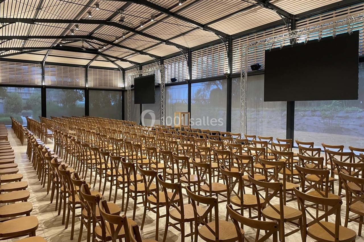 Salle de conférence vide avec des rangées de chaises en bois, un pupitre central et deux écrans noirs suspendus.