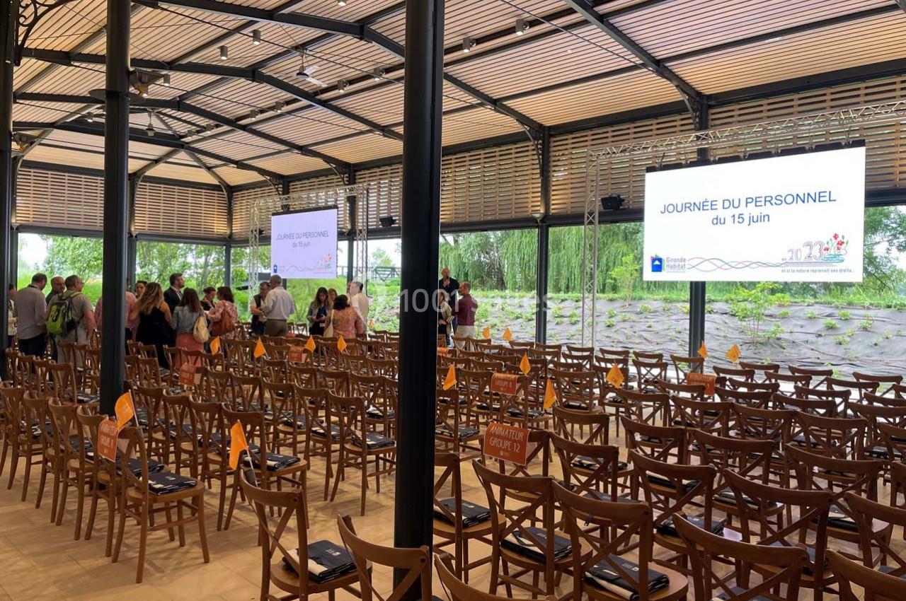 Salle avec des chaises en bois, écrans affichant ’Journée du personnel du 15 juin’ et participants debout au fond.