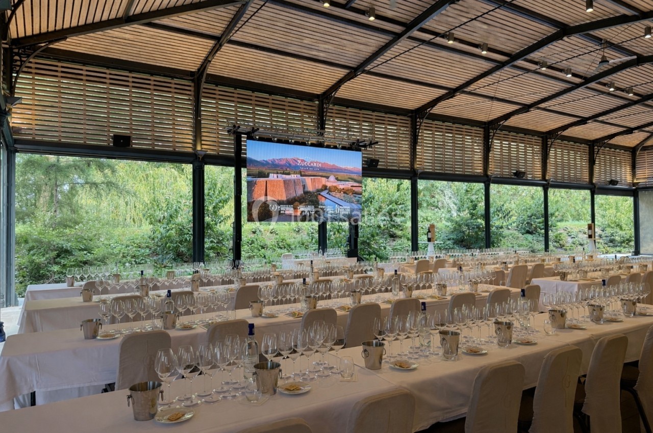 Salle lumineuse avec de grandes baies vitrées, tables dressées pour une dégustation de vins, écran affichant un paysage.