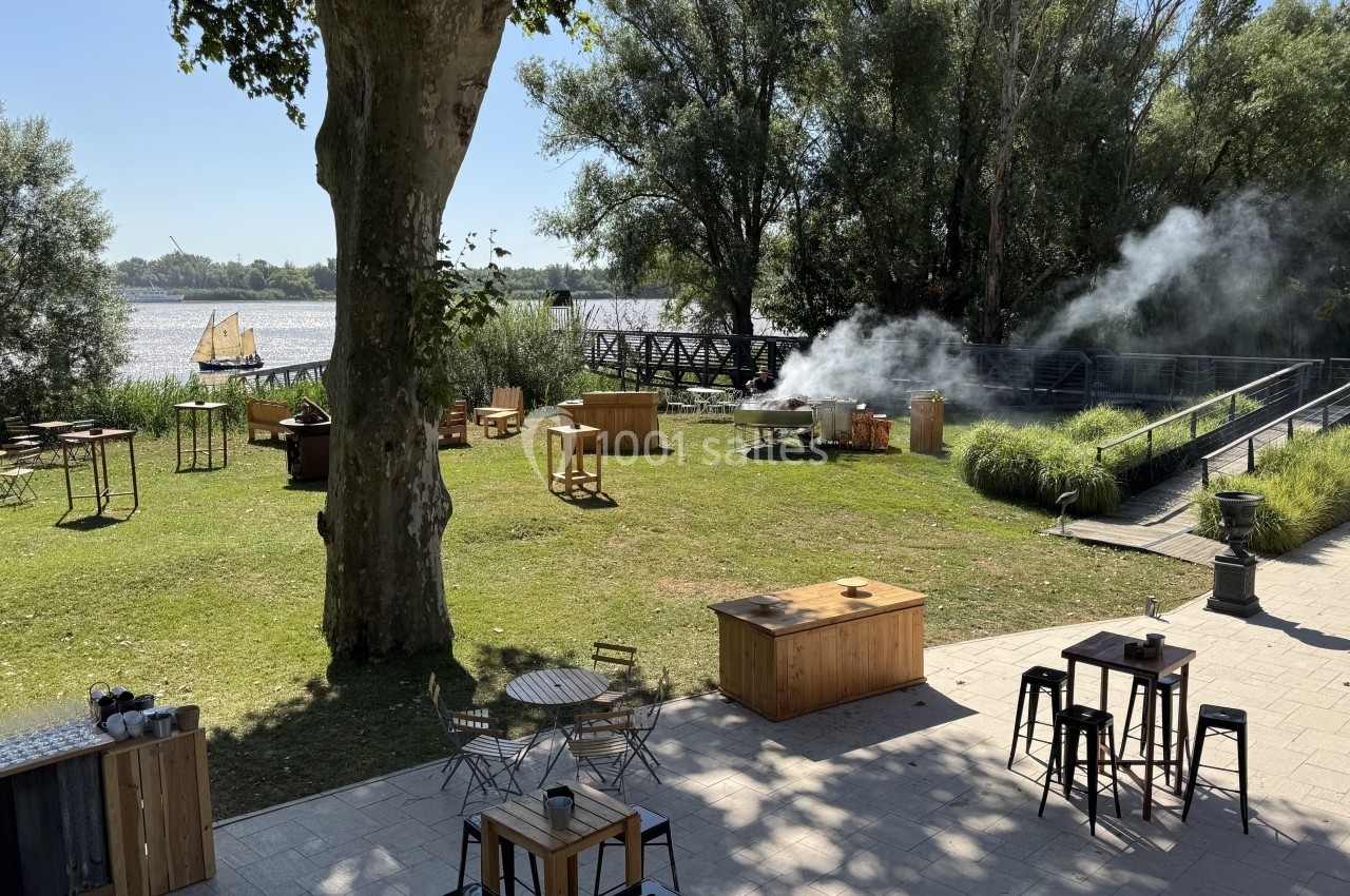 Espace extérieur verdoyant avec tables, chaises et fumée près d'un barbecue, vue sur un fleuve et un voilier au loin.