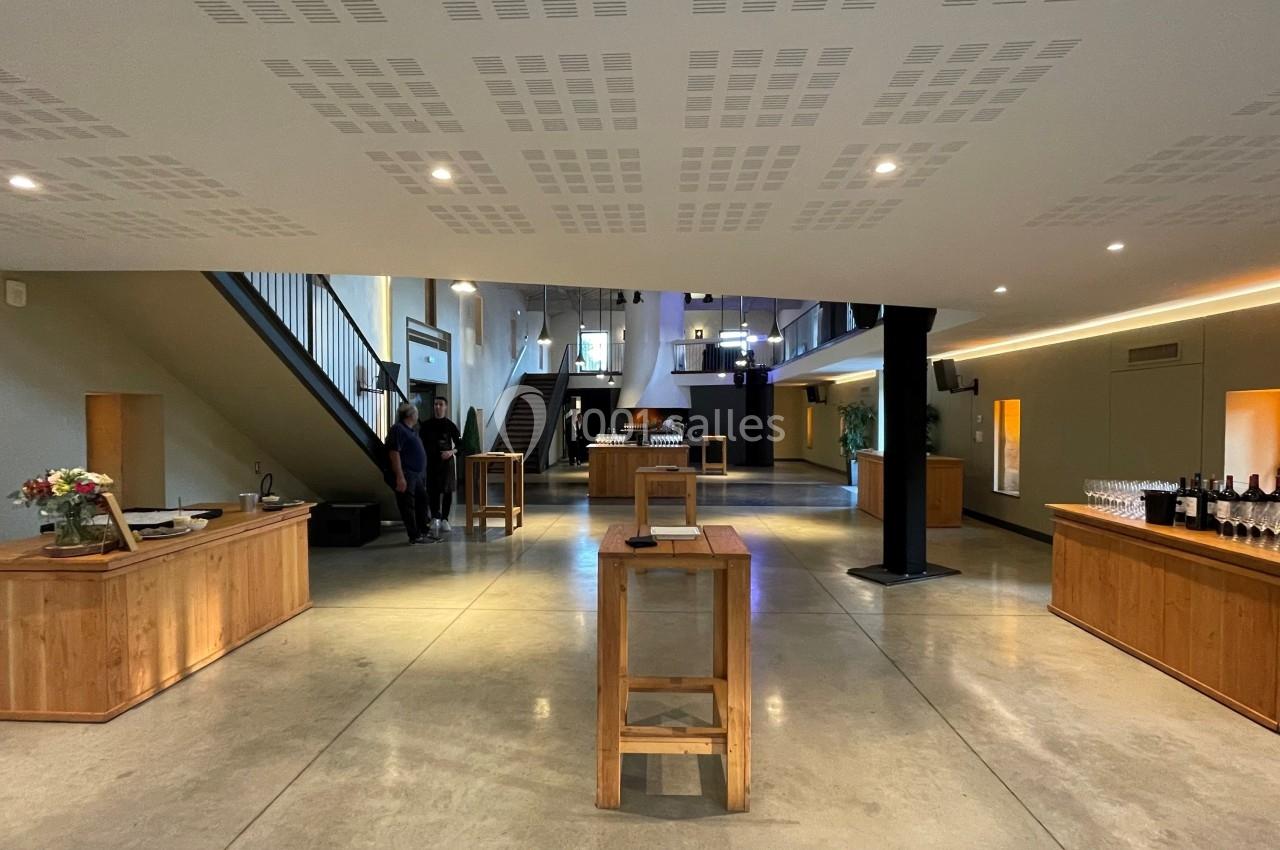 Salle lumineuse avec mobilier en bois, tables hautes, escalier et espace aménagé pour une réception ou un événement.