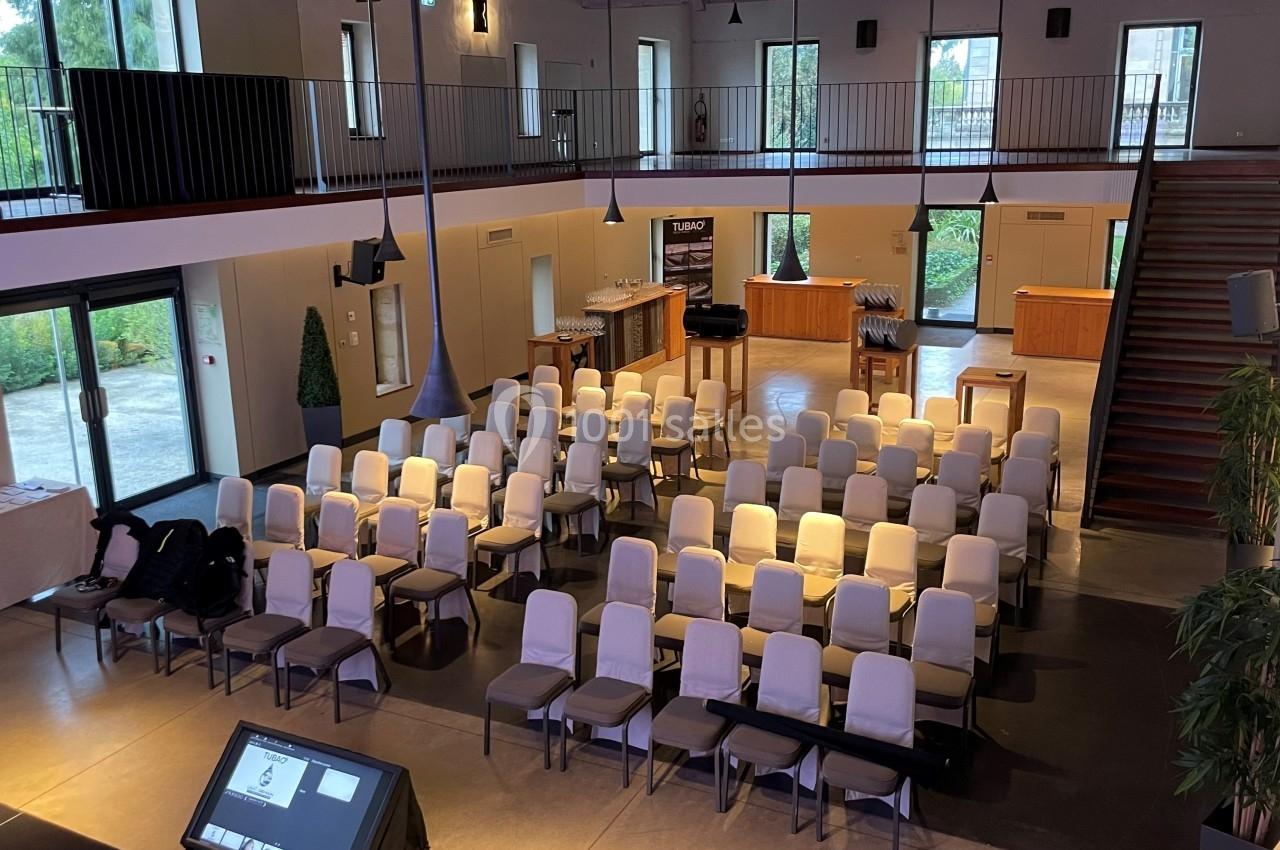Salle de conférence lumineuse avec des rangées de chaises alignées, un écran au premier plan et un balcon à l'étage.
