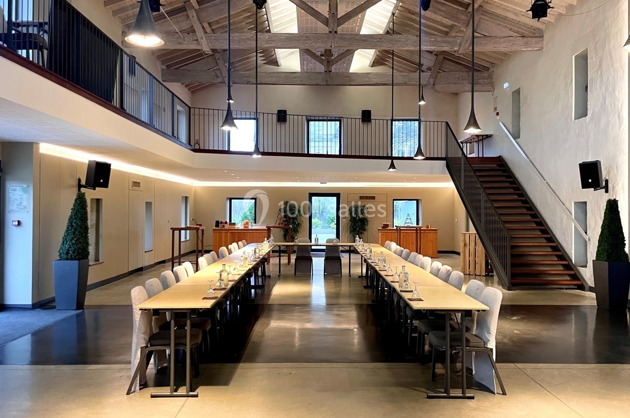 Grande salle lumineuse avec tables alignées en U, chaises, escalier en bois et plafond avec poutres apparentes.