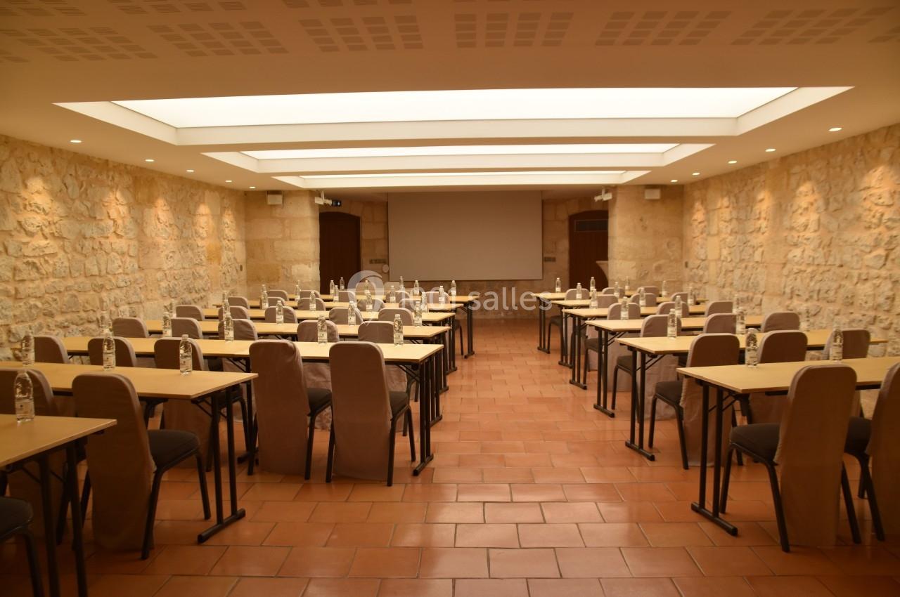 Salle de conférence avec des murs en pierre, des tables et des chaises alignées, éclairée par des plafonniers.