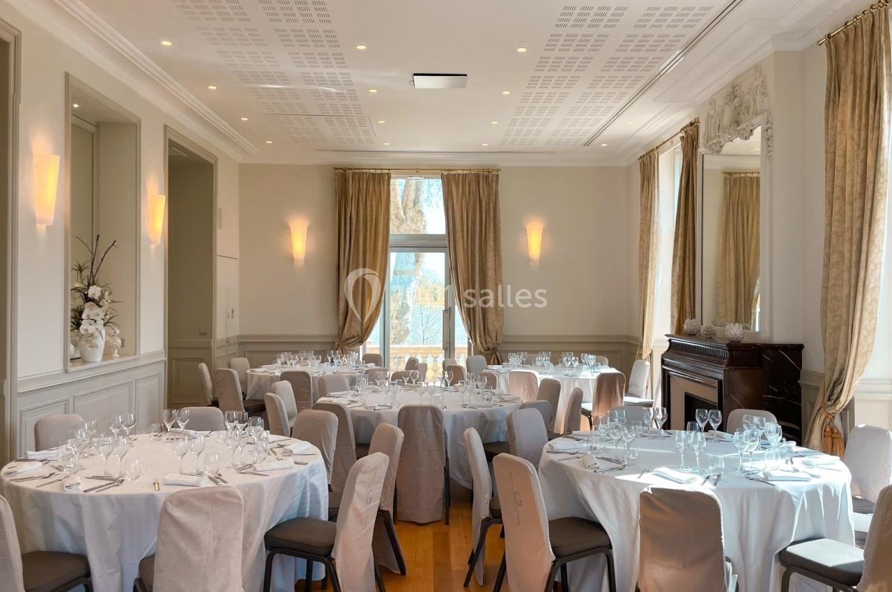 Salle lumineuse avec tables rondes dressées pour un événement, rideaux beiges et cheminée décorative en arrière-plan.