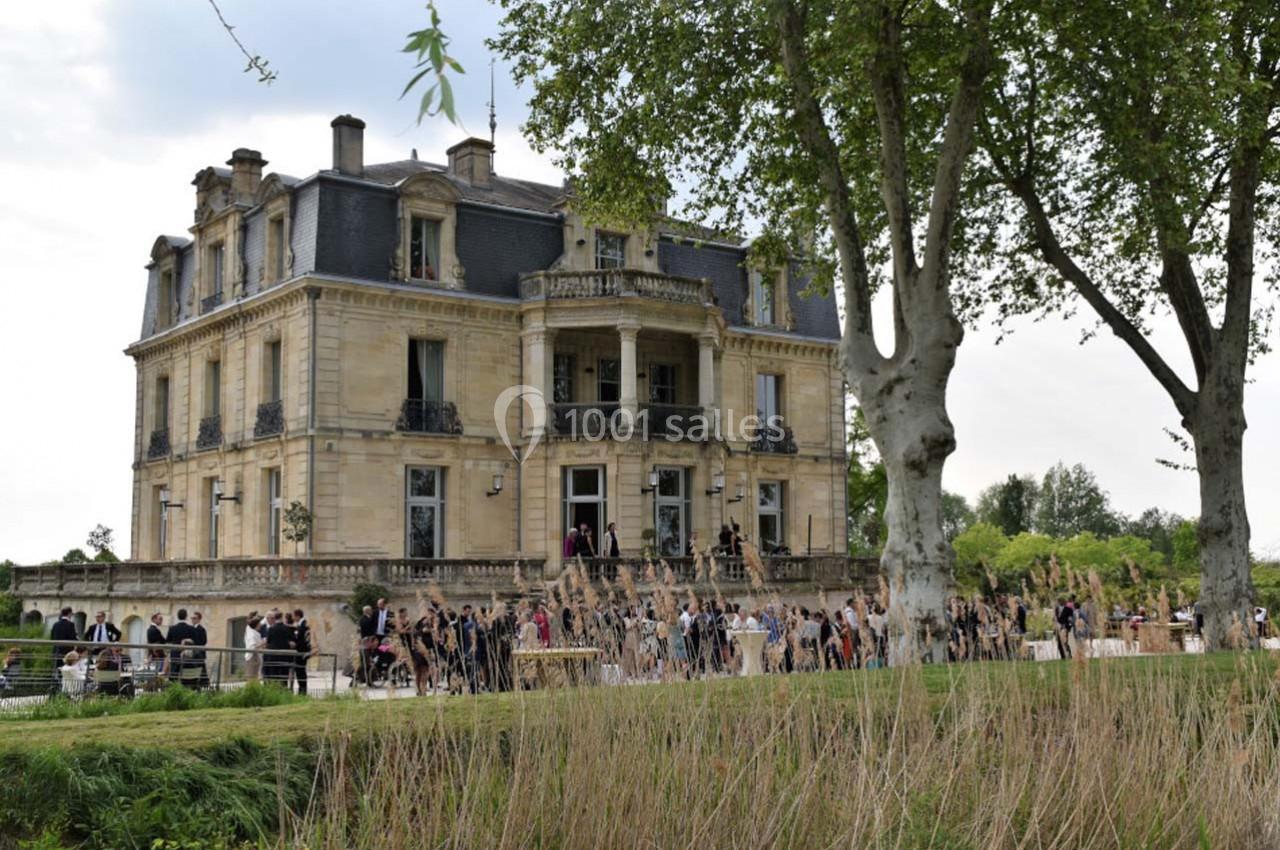 Façade d'un château en pierre entouré d'arbres, avec des personnes rassemblées sur l'esplanade devant.