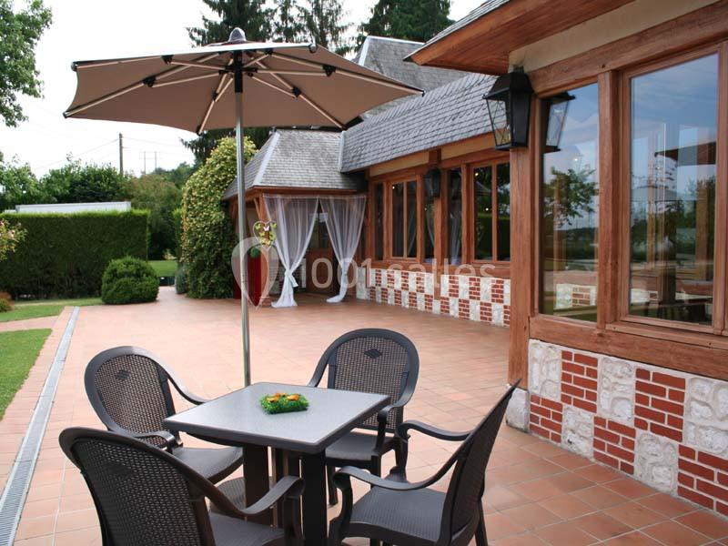Table et chaises en terrasse pavée, avec parasol, près d'un bâtiment en briques et bois aux grandes fenêtres.