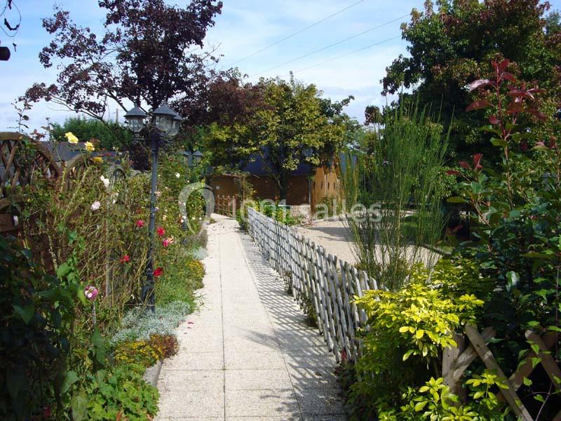 Allée de jardin bordée de fleurs colorées et de haies, menant à une petite cabane entourée d'arbres.