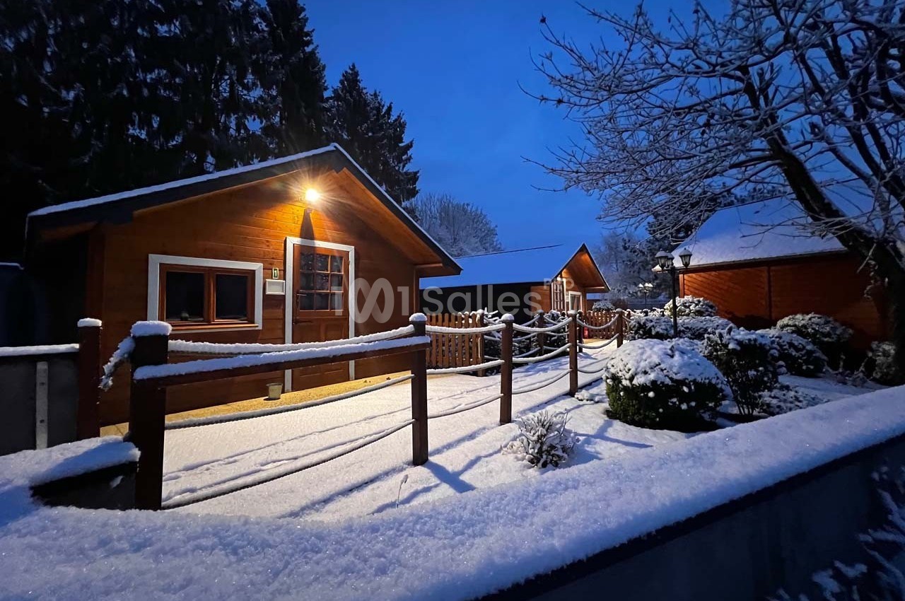Chalets en bois éclairés par une lumière chaude, entourés de neige et d'arbres sous un ciel crépusculaire.