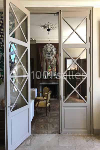 Porte vitrée à croisillons ouverte sur un salon lumineux avec des fauteuils anciens et un lustre décoratif.