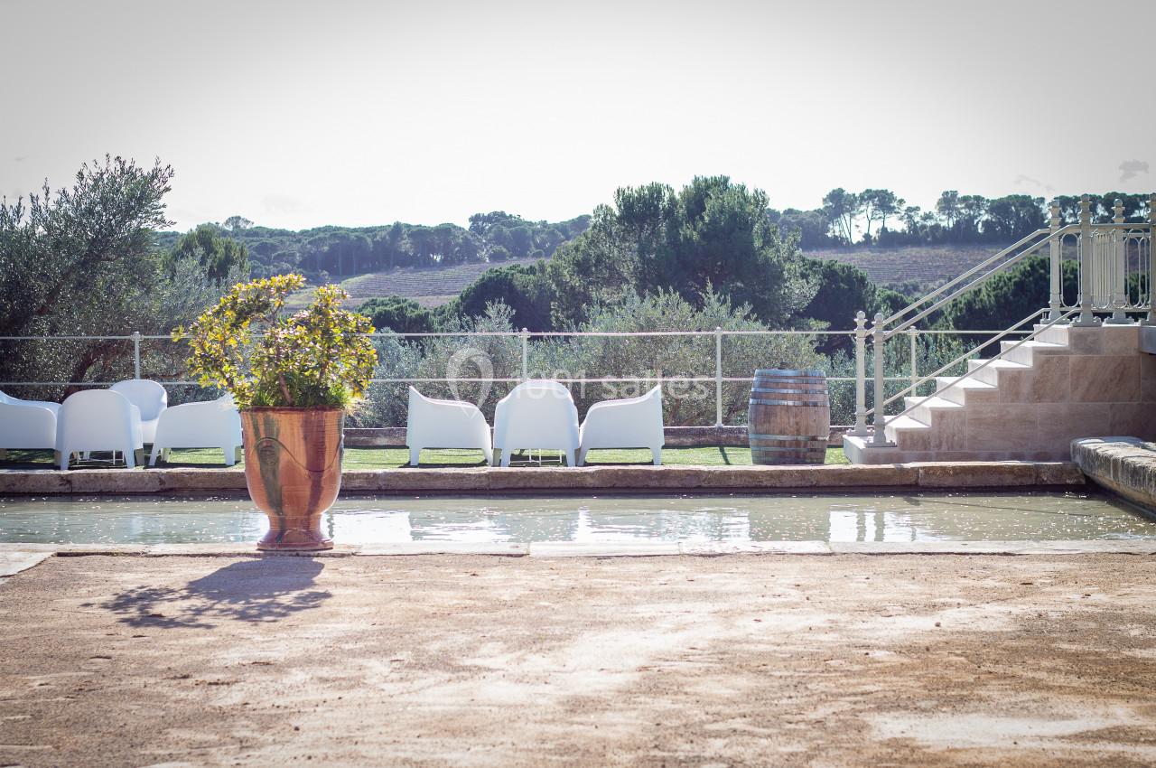Terrasse avec bassin, pot de fleurs, chaises blanches et vue sur un paysage arboré sous un ciel clair.
