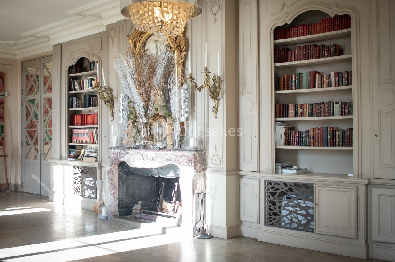 Bibliothèque lumineuse avec cheminée décorative, étagères remplies de livres et lustre en cristal.