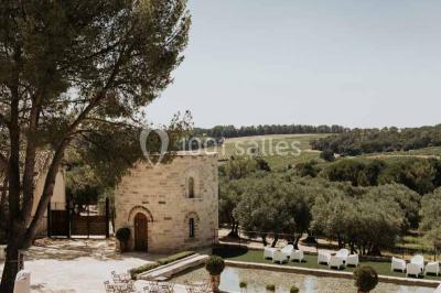 Tour en pierre entourée de végétation, avec bassin d'eau, chaises et tables en métal sur une terrasse ensoleillée.