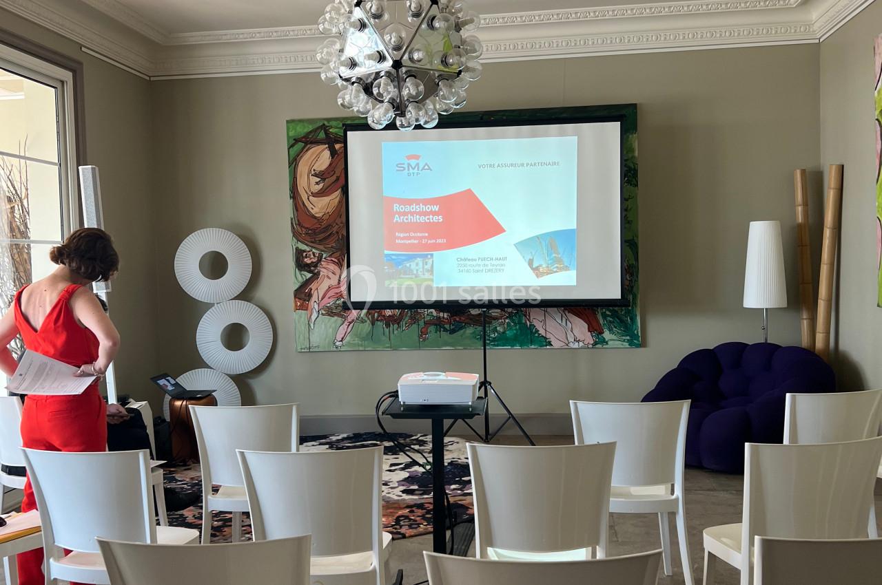 Salle de conférence lumineuse avec des chaises blanches alignées, un projecteur allumé et une présentation affichée.