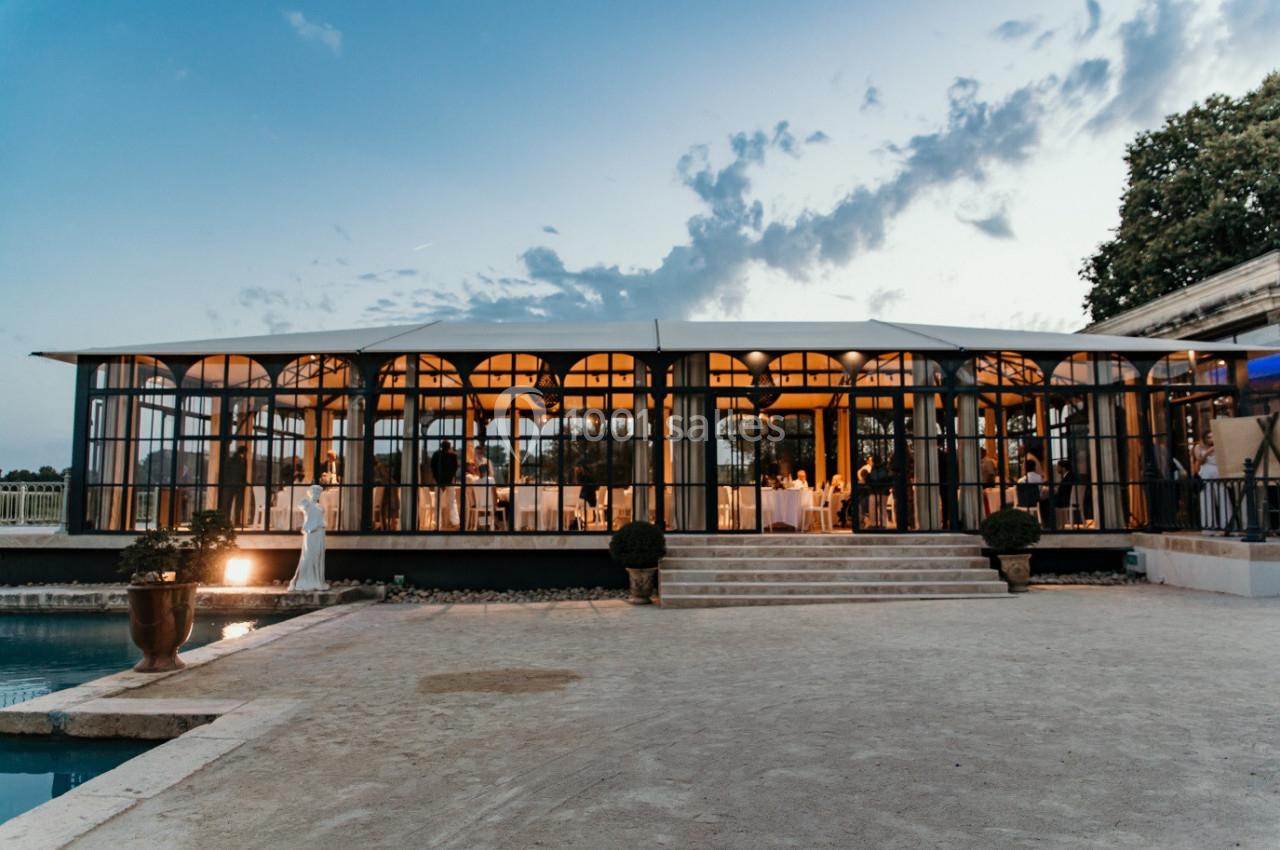 Salle vitrée éclairée en soirée, entourée d'un jardin avec piscine au premier plan.