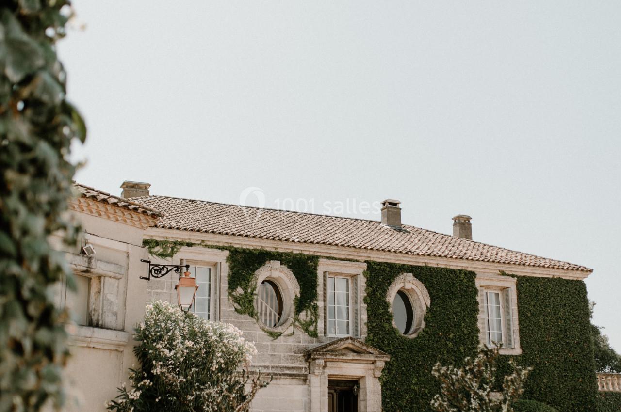 Façade d'une maison en pierre recouverte de lierre, avec des fenêtres rondes et des plantes en pots devant l'entrée.