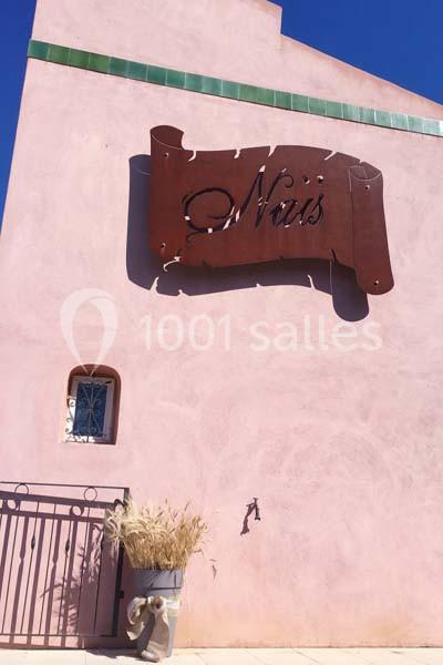 Façade rose avec une enseigne métallique ’Naïs’, une petite fenêtre, un robinet mural et un pot de plantes sèches.