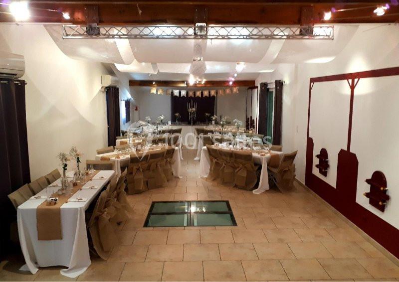 Salle de réception décorée avec des tables dressées, nappes blanches et chaises couvertes, sous un plafond en poutres…