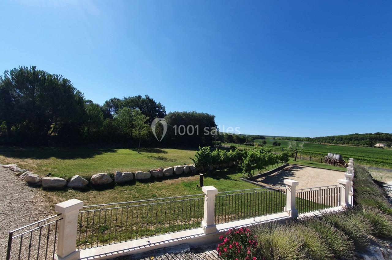 Vue d'un jardin avec pelouse, arbres, allée en gravier, clôture et paysage de campagne sous un ciel bleu.