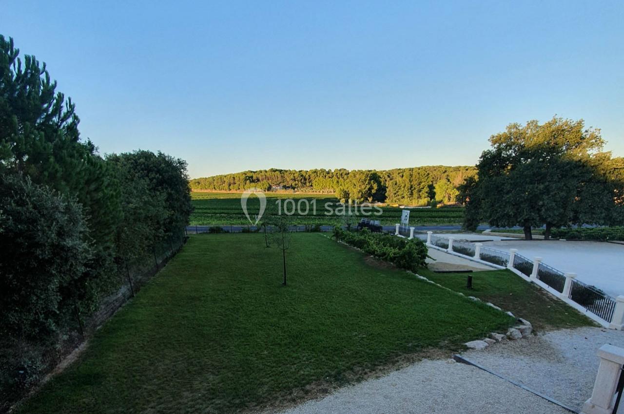 Vue d'un jardin avec pelouse, arbres et allée gravillonnée, donnant sur des vignes et une forêt à l'horizon.