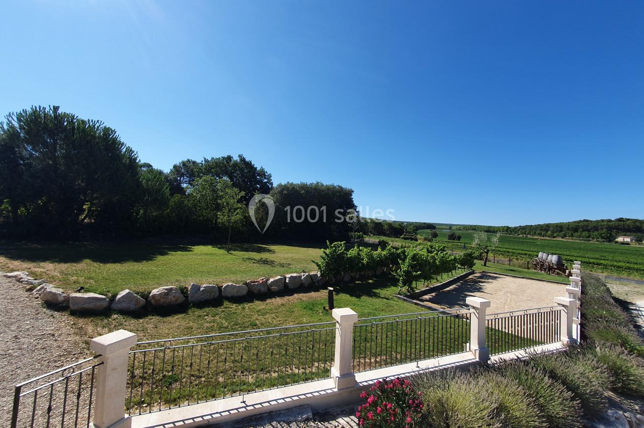 Vue d'un jardin avec pelouse, arbres, allée gravillonnée, clôture et paysage rural sous un ciel bleu dégagé.