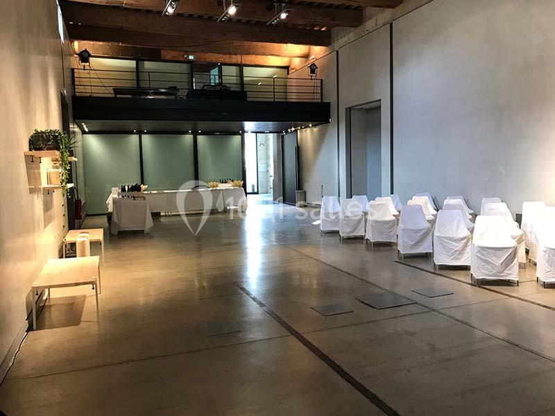 Location salle Avignon (Vaucluse) - Le Grenier à Sel #6 Salle lumineuse avec sol en béton, chaises blanches alignées, tables dressées au fond et mezzanine en bois.