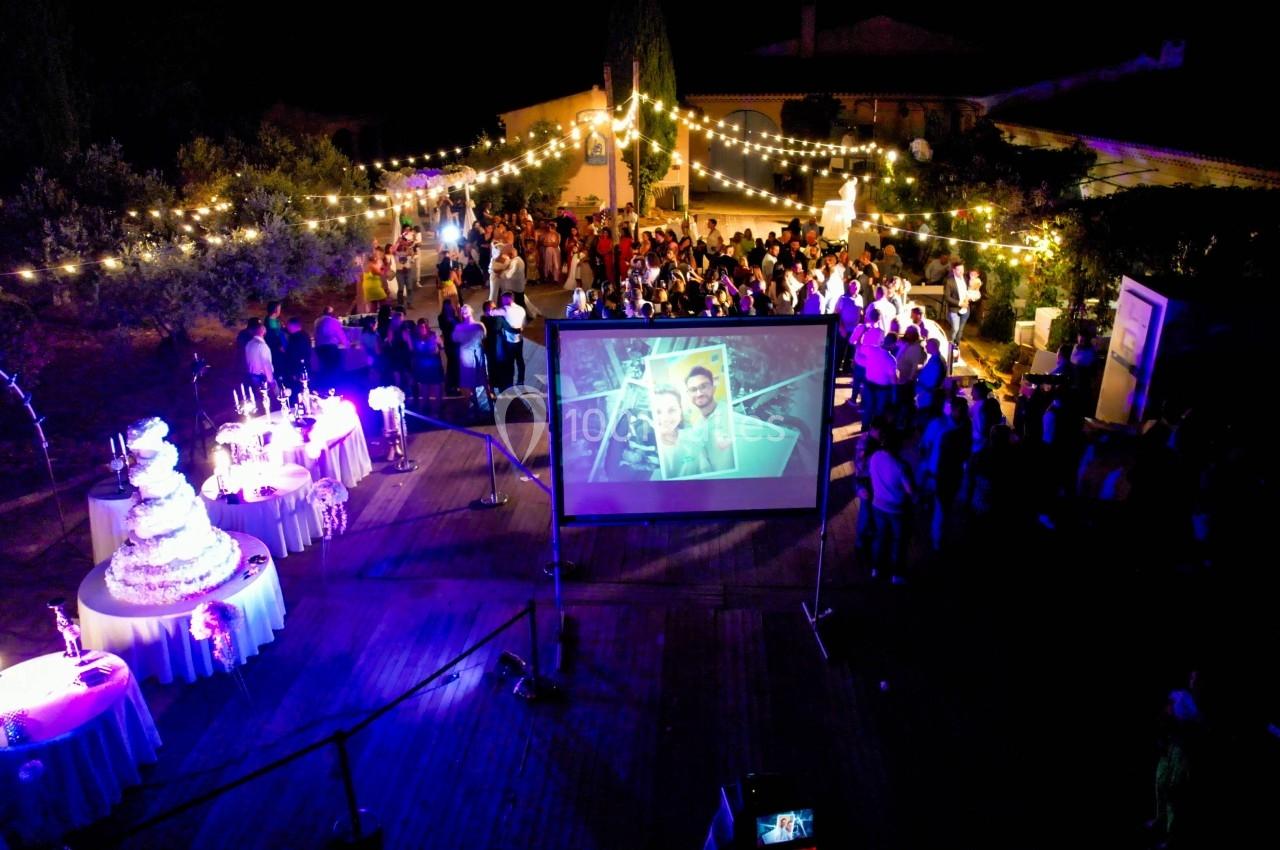 Vue nocturne d'une fête en plein air avec des guirlandes lumineuses, un écran de projection et des invités rassemblés.