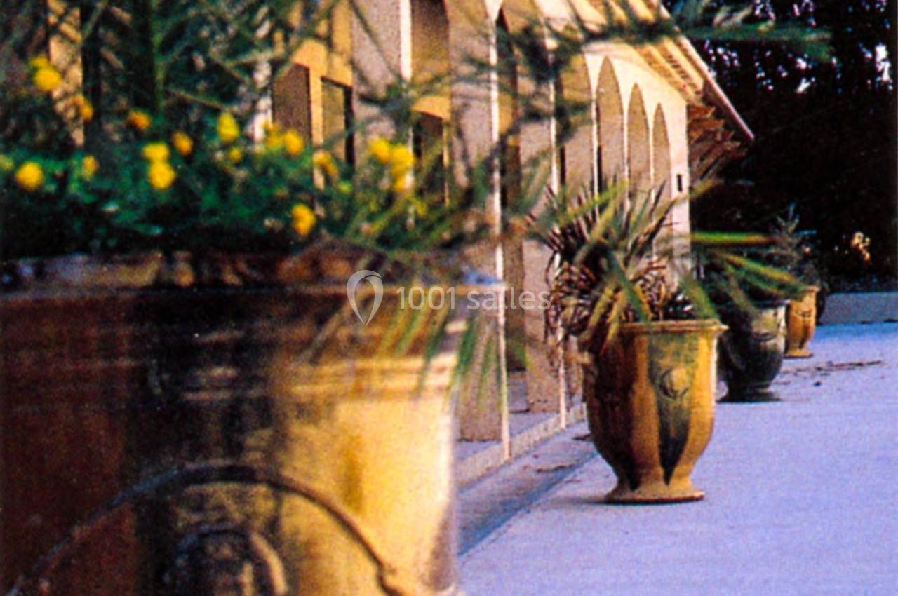 Alignement de grands pots en terre cuite avec des plantes, devant un bâtiment à arcades et une allée bordée d'arbres.
