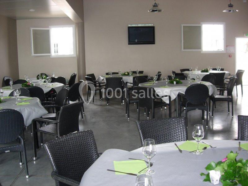 Salle de réception avec tables rondes dressées, nappes blanches, serviettes vertes et chaises en osier noir.
