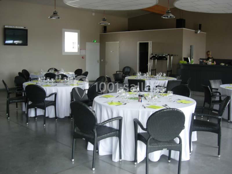 Salle de réception avec tables rondes dressées, nappes blanches, chaises en osier noir et un bar en arrière-plan.