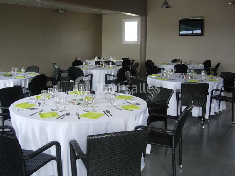 Salle de réception avec tables rondes dressées, nappes blanches, chaises noires et serviettes vertes.
