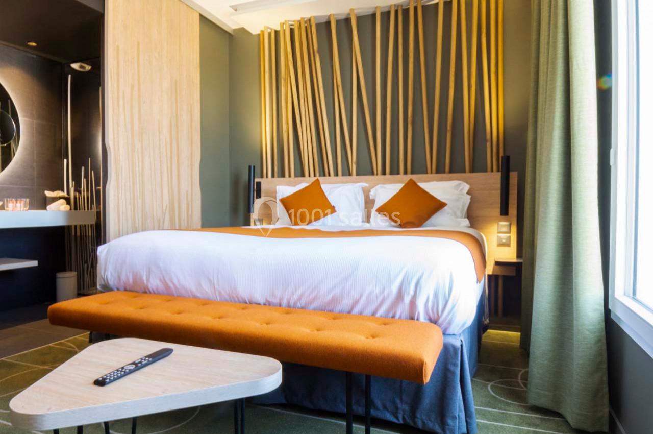 Location salle Montpellier (Hérault) - Hôtel Belaroïa #8 Chambre d'hôtel moderne avec lit double, coussins orange, décoration en bois et lumière naturelle provenant d'une fenêtre.