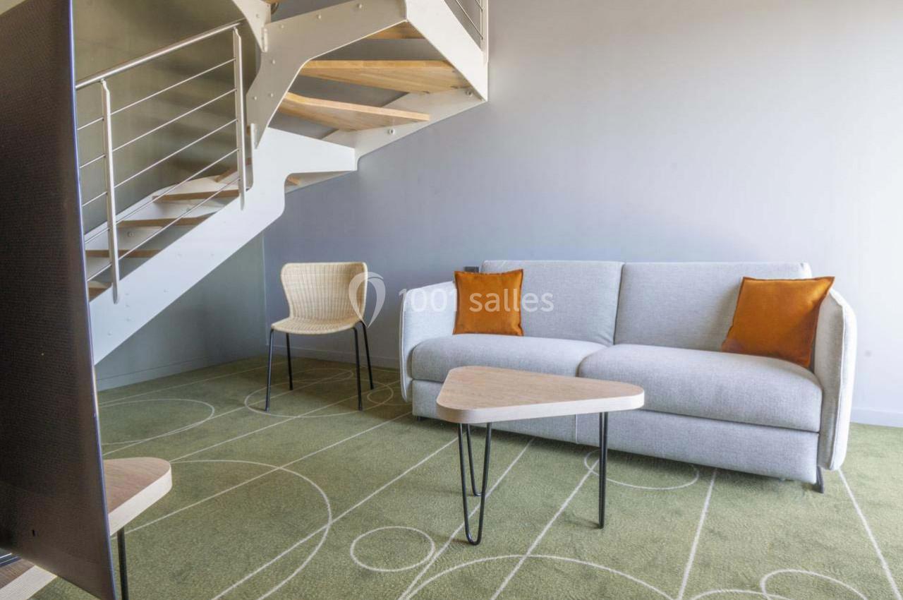 Location salle Montpellier (Hérault) - Hôtel Belaroïa #14 Salon moderne avec canapé gris, coussins orange, chaise en osier, table basse triangulaire et escalier en métal.