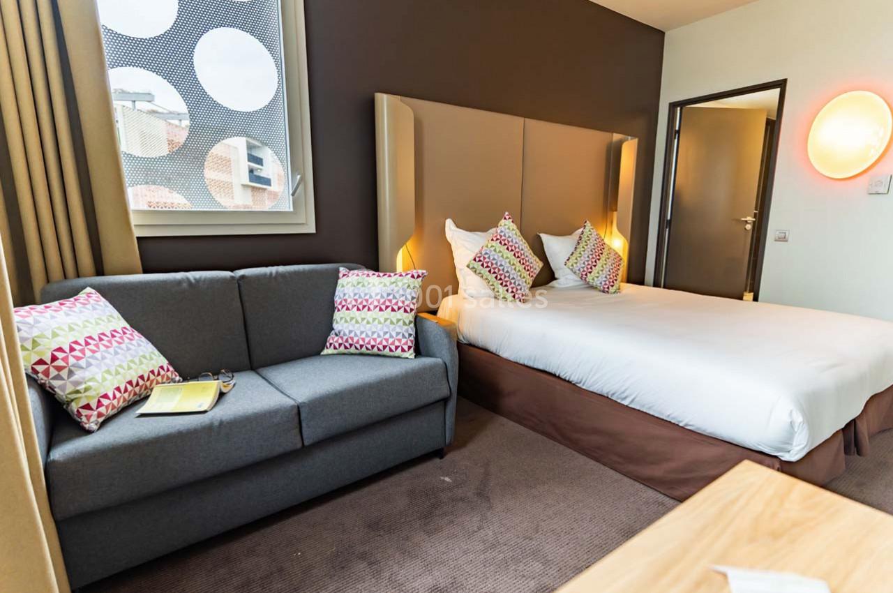 Location salle Montpellier (Hérault) - Hôtel Belaroïa #9 Chambre d'hôtel avec lit double, canapé gris, coussins colorés, fenêtre et éclairage mural.