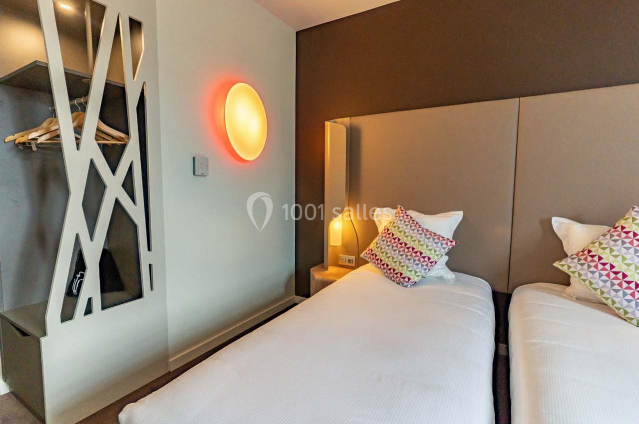 Location salle Montpellier (Hérault) - Hôtel Belaroïa #12 Deux lits simples avec têtes de lit rembourrées, coussins colorés, éclairage mural et espace de rangement ouvert.