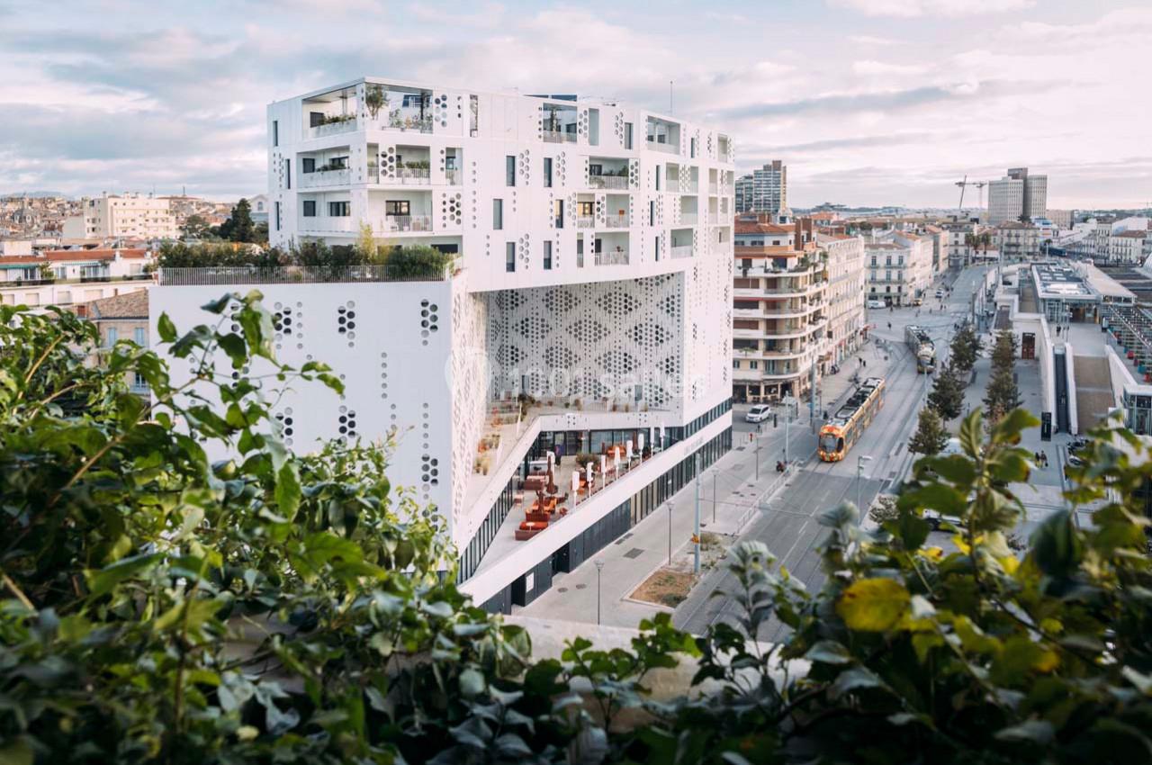 Location salle Montpellier (Hérault) - Hôtel Belaroïa #3 Immeuble moderne blanc avec motifs perforés, entouré de bâtiments urbains et d'une rue avec tramway.