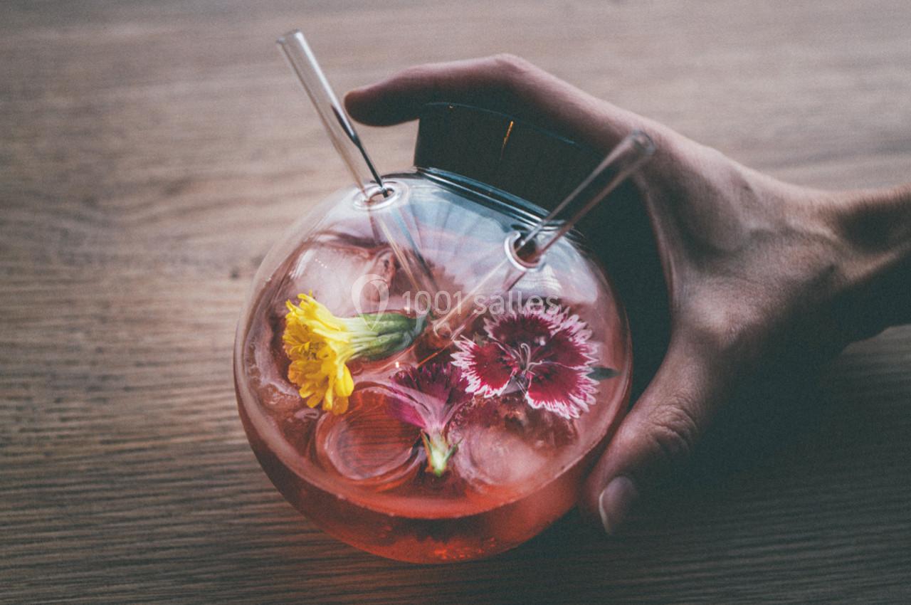 Location salle Montpellier (Hérault) - Hôtel Belaroïa #7 Main tenant un verre rond rempli d'une boisson rose, décorée de glaçons et de fleurs colorées, sur une table en bois.