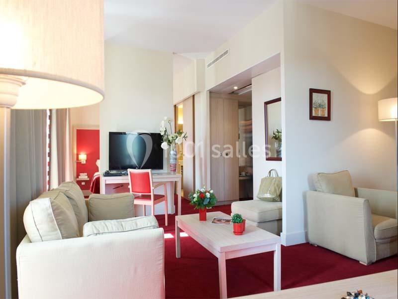 Salon lumineux avec canapé beige, fauteuil, table basse, bureau, télévision et décoration florale sur moquette rouge.