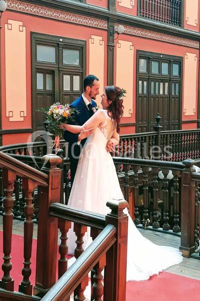 Un couple en tenue de mariage pose sur un escalier en bois dans un bâtiment au style architectural ancien.