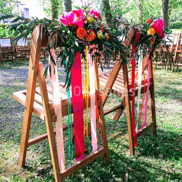 Deux chaises en bois décorées de rubans colorés et de bouquets de fleurs vives dans un cadre extérieur verdoyant.