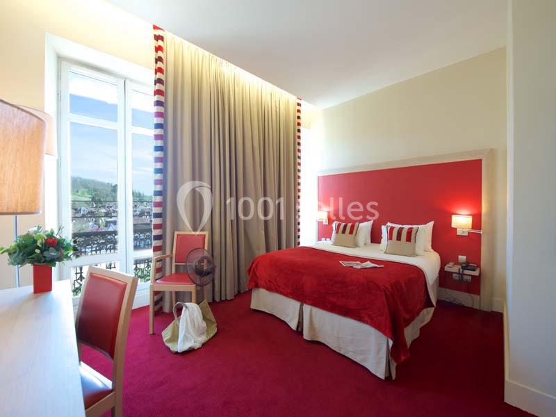 Chambre lumineuse avec lit double, tête de lit rouge, rideaux beiges, bureau en bois et vue sur un paysage extérieur.