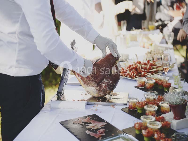 Un serveur découpe du jambon sur un buffet garni de verrines et brochettes lors d'un événement en extérieur.