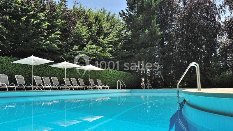 Piscine extérieure entourée de chaises longues, parasols blancs et haies verdoyantes sous un ciel dégagé.