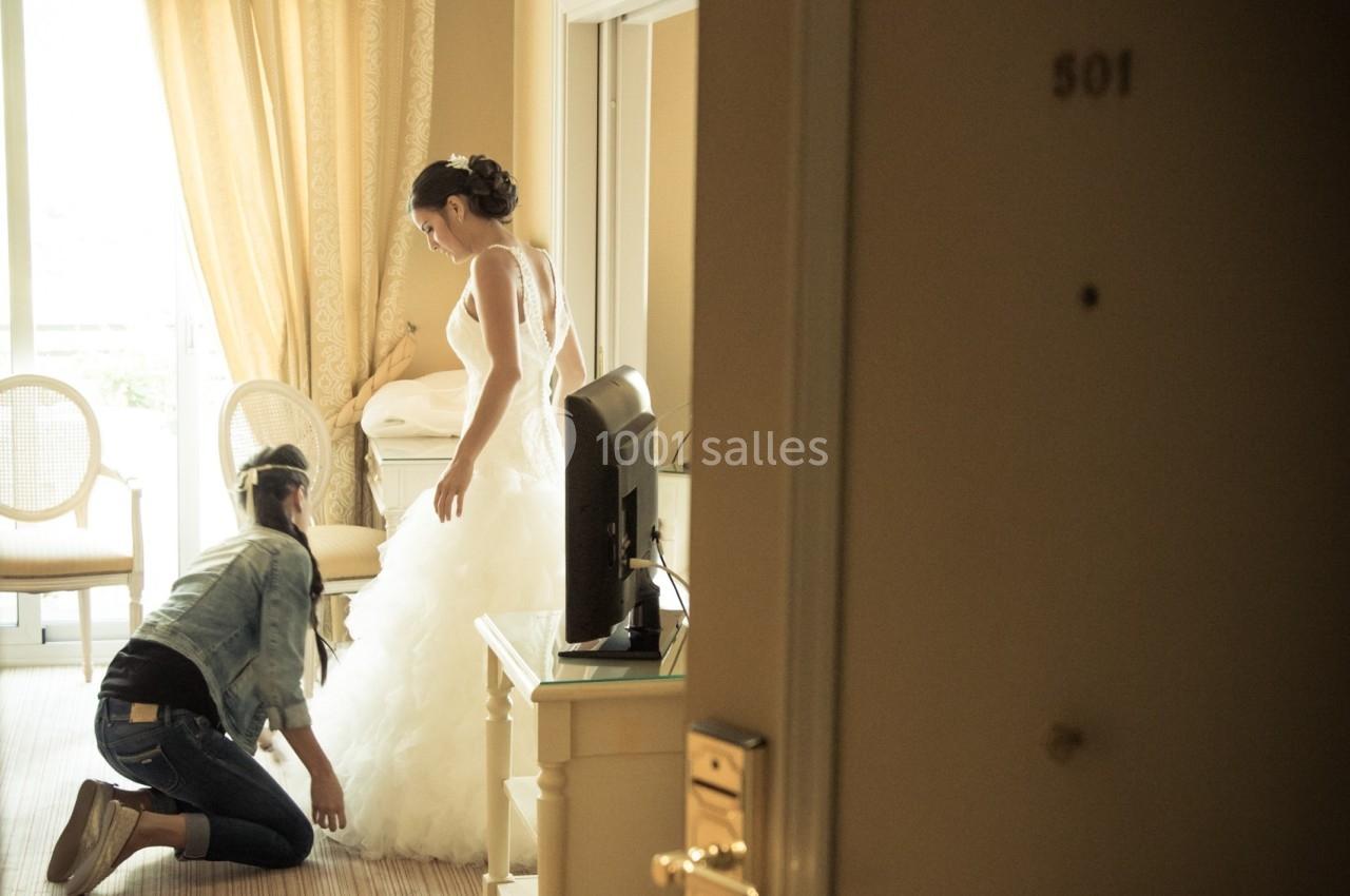 Une femme ajuste la robe de mariée d'une autre femme dans une chambre d'hôtel lumineuse.