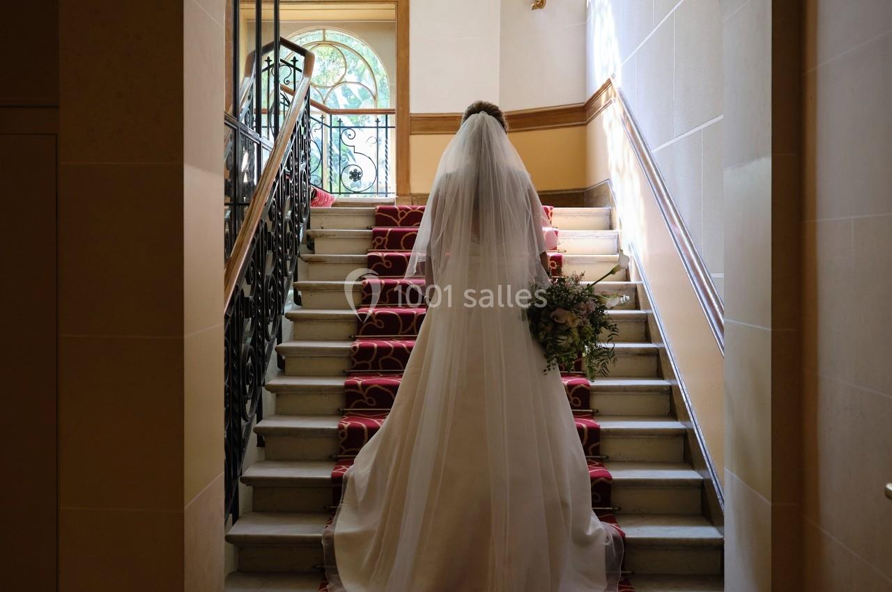 Une mariée en robe blanche monte un escalier décoré de tapis rouge dans un intérieur lumineux.