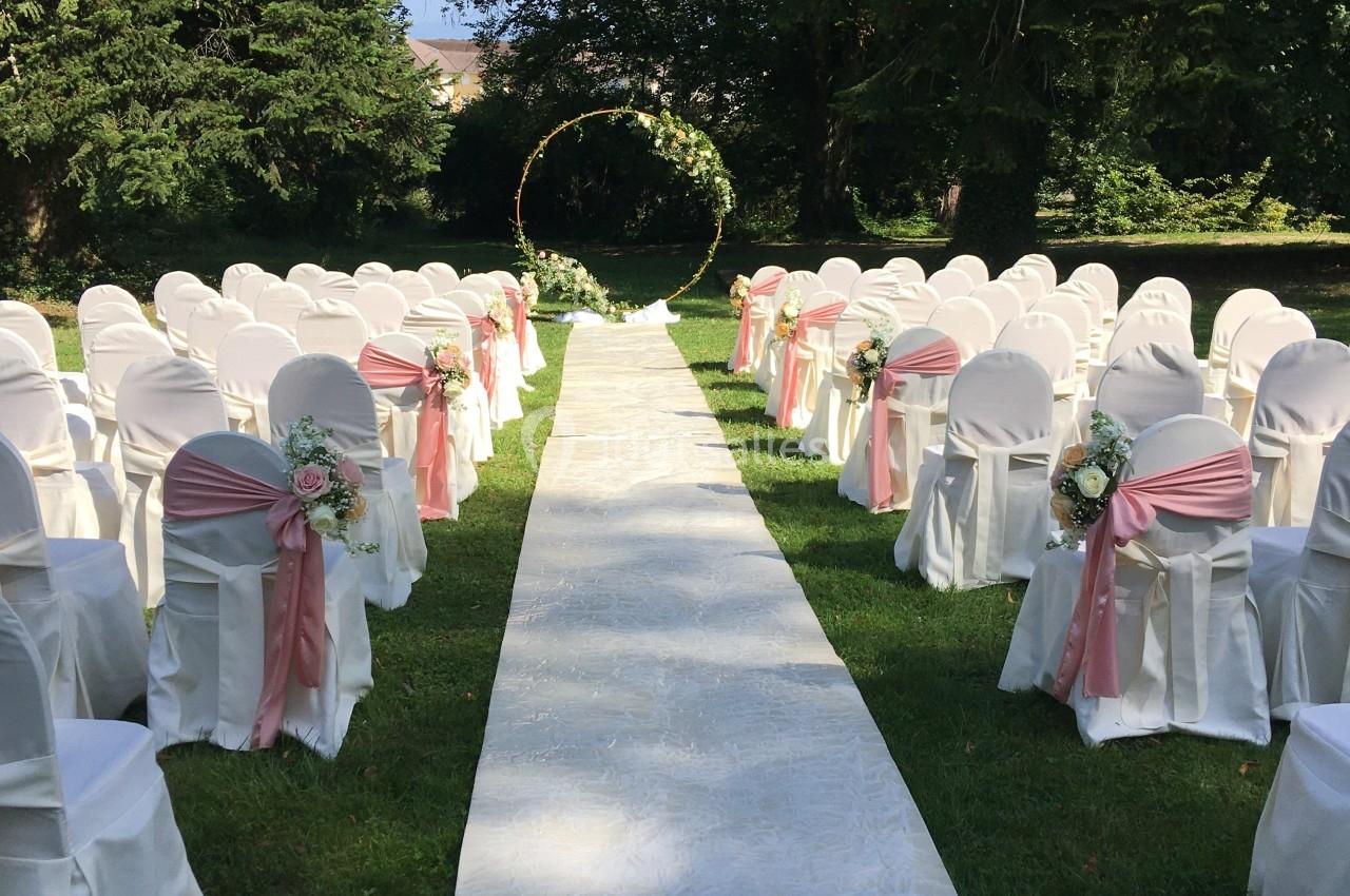 Allée centrale bordée de chaises blanches décorées de rubans roses, menant à une arche florale dans un jardin verdoyant.