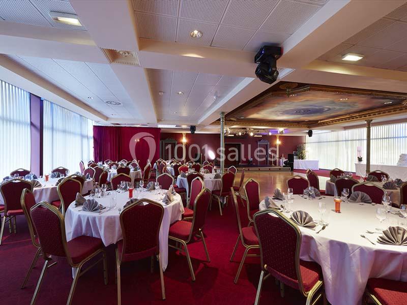 Salle de réception avec tables rondes dressées, nappes blanches, serviettes pliées et chaises rouges.